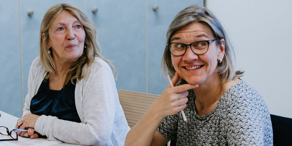 Zwei Frauen sitzen während Fortbildungen an einem Tisch in einem Büro oder Klassenzimmer. Eine Frau mit langen Haaren blickt zur Seite, während die andere, die eine Brille trägt, lächelt und sich ans Kinn fasst. Auf dem Tisch vor ihnen liegen Papiere.